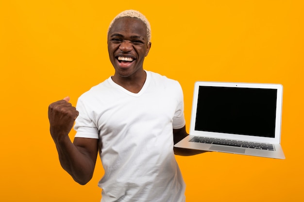 Joyful african man waving his hands holding a laptop with a mockup on a yellow background