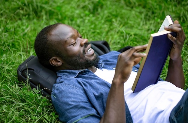 즐거운 아프리카계 미국인 학생은 캠퍼스 공원 잔디밭에 누워 책을 읽습니다.