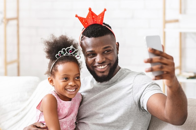 Joyful afro family father and preschool daughter having fun at home, taking selfie, wearing crowns