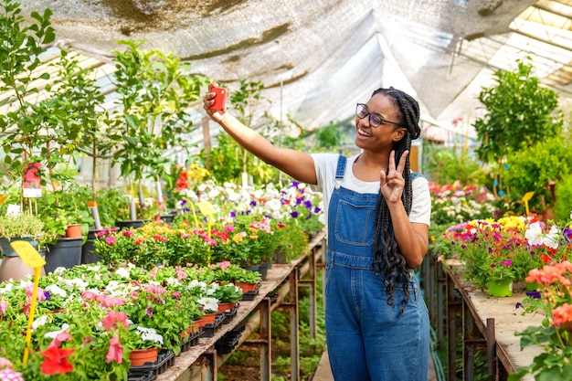 즐거운 아프리카계 미국인 식물 가게 직원이 다채로운 온실에서 평화로운 셀피를 찍습니다.