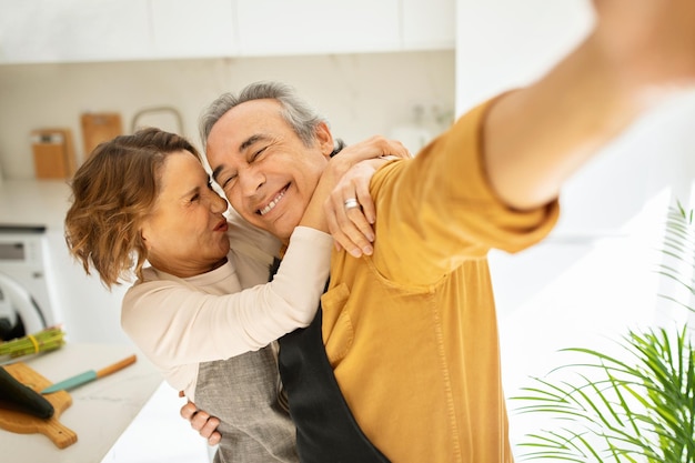 Joyful aged married couple making selfie grimacing and hugging in kitchen interior taking photos of