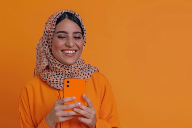 20대 후반의 즐거운 아랍 여성이 긍정과 열정을 방출하는 활기찬 오렌지색 배경에서 스마트폰 앱을 추천하면서 따뜻하게 미소 짓고 있습니다.
