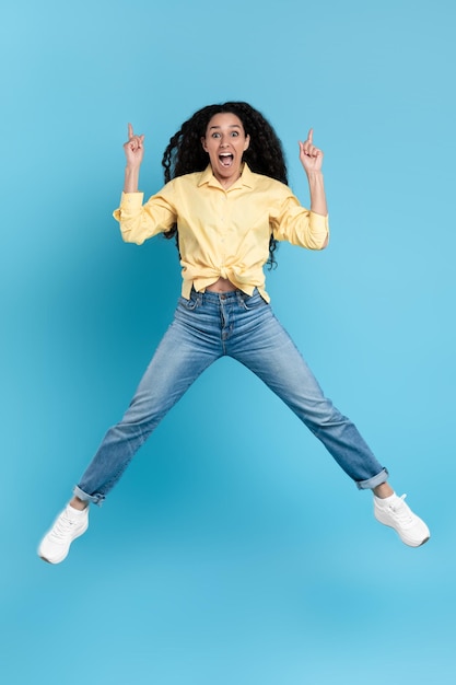 Photo joyful arabic female jumping pointing fingers up over blue background