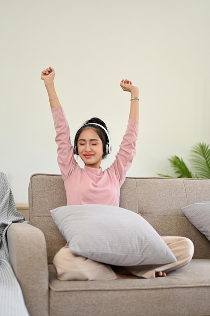 Joyful Asian girl listening music on headphones raising hands and relaxing on her sofa