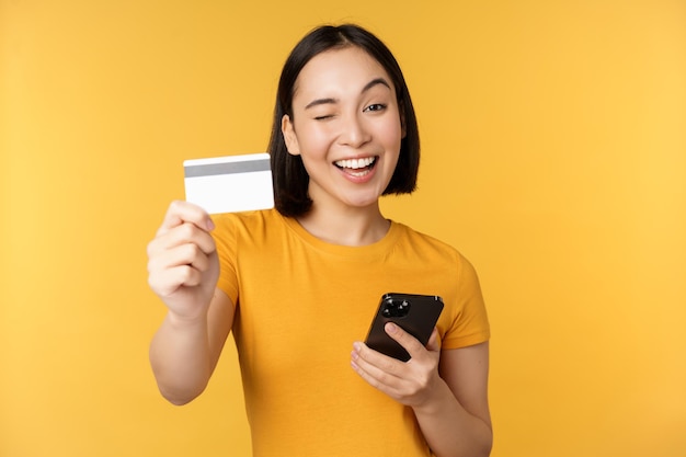 Радостная азиатская девушка улыбается, показывая кредитную карту и смартфон, рекомендуя мобильный телефон, стоя на желтом фоне