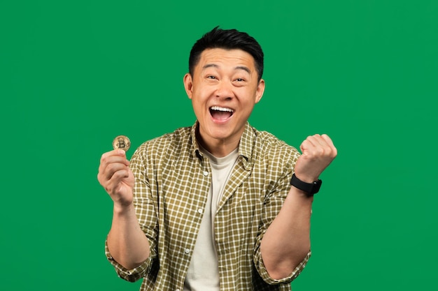 Joyful asian mature man showing yes gesture and holding golden bitcoin in his hand posing over green