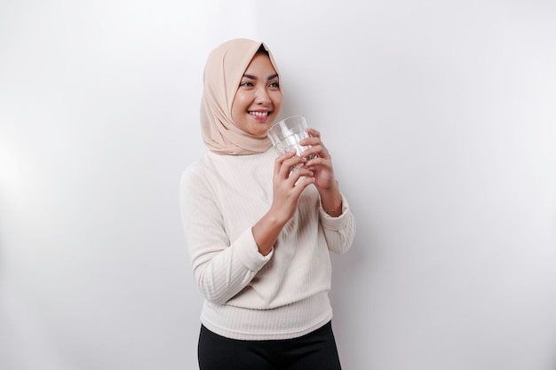 Joyful Asian Muslim woman wearing headscarf is drinking a glass of water isolated on white background