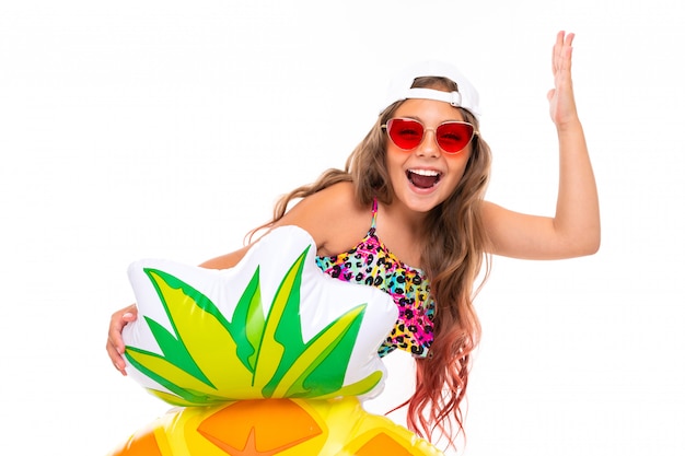 Joyful attractive girl in a swimsuit and a baseball cap with a rubber ring on a white wall