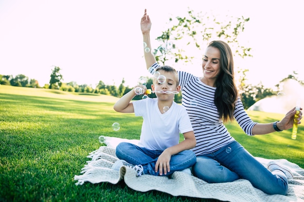 うれしそうな魅力的な母親は、彼女の小さなかわいい息子と一緒に座って、夏の緑豊かな公園で日没時にバブルブロワーで遊んでいます