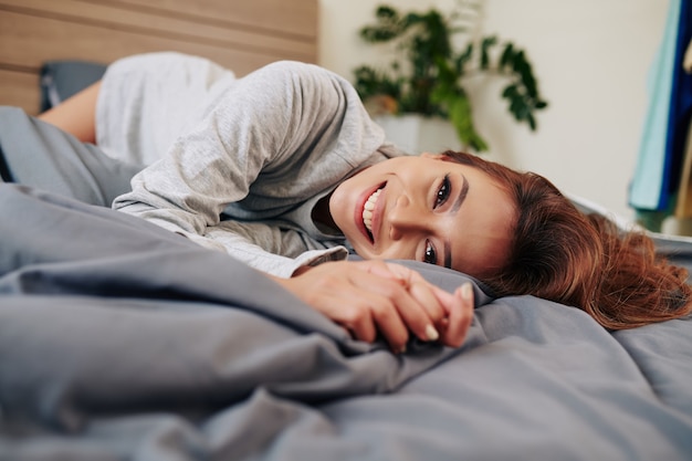 즐거운 매력적인 젊은 아시아 여성이 집에서 편안한 침대에서 휴식을 취하고 카메라를 바라보고 있습니다.