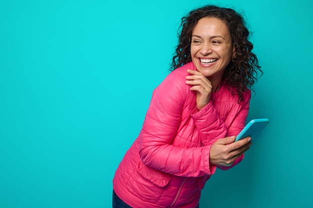 Joyful awesome attractive pretty woman with curly hairstyle, wearing bright pink jacket, laughs holding smartphone in her hand and looking through her shoulders at a copy ad space of blue background