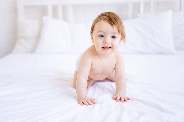 6 개월 된 기쁜 아기 소녀는 기저귀를 입고 침대에 기어서 웃고, 집에서 면화 침대에 작은 아이는 보살과 위생의 개념을 가지고 있습니다.