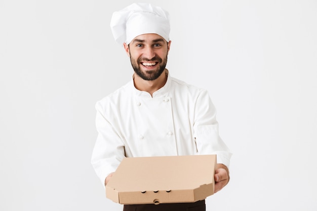 joyful baker man in cook uniform smiling and holding pizza box isolated over white wall