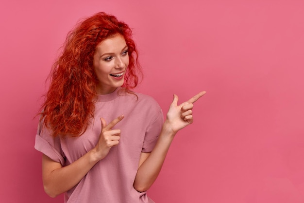 Joyful beautiful red-haired young girl student points finger to left on pink background