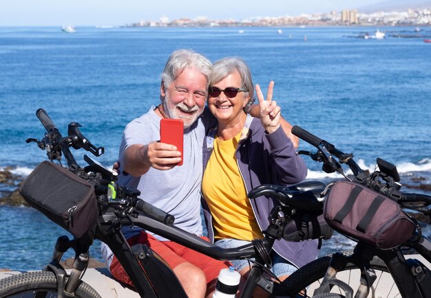 海の地平線、自転車の近くの屋外遠足で電話を使用してうれしそうな美しい年配のカップル。健康的なライフスタイルを楽しむアクティブな退職者