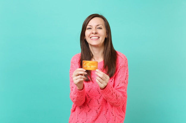 Joyful beautiful young girl in knitted pink sweater looking camera, hold in hand credit card isolated on blue turquoise wall background studio portrait. People lifestyle concept. Mock up copy space.