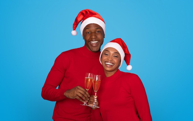 산타 모자를 입은 즐거운 흑인 커플이 스튜디오에서 푸른 배경에  ⁇ 페인 컵을  ⁇ 들여 크리스마스 휴가 시즌을 축하하고 카메라에 미소 짓고 따뜻한 축제 소원을 주고받고 있습니다.