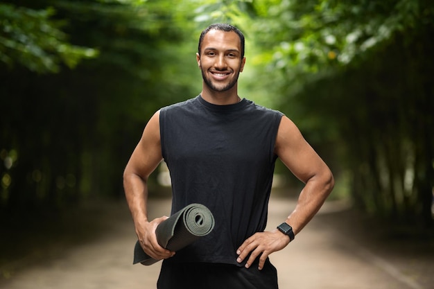 Joyful black guy having workout at public park holding mat