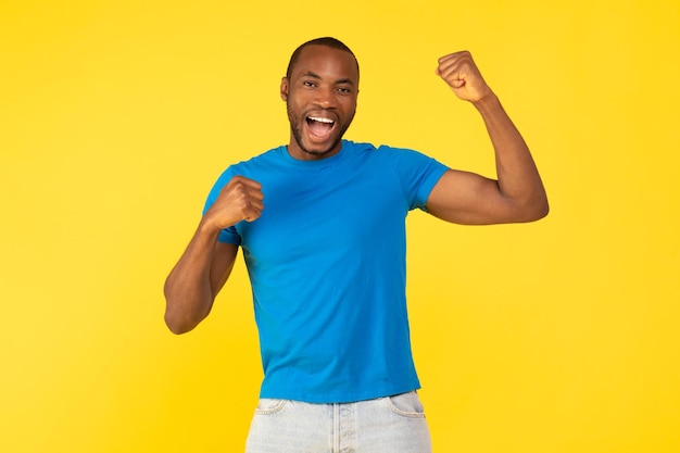 Joyful black male shaking fists celebrating success over yellow background