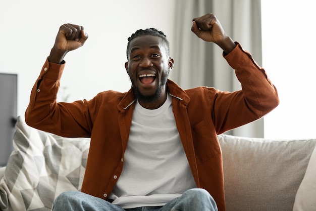 Joyful black man cheering for sports team on television gesturing YES and shouting sitting on sofa at home