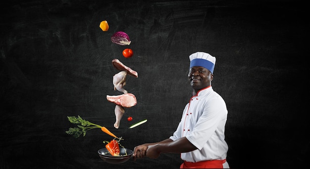 Joyful black man wearing an apron and cooking in action, chalkboard background. Mixed media