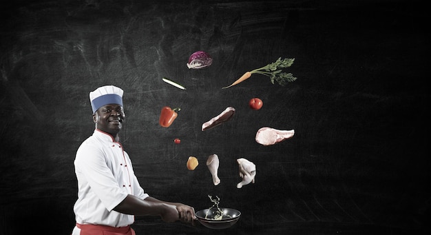 Joyful black man wearing an apron and cooking in action, chalkboard background. Mixed media