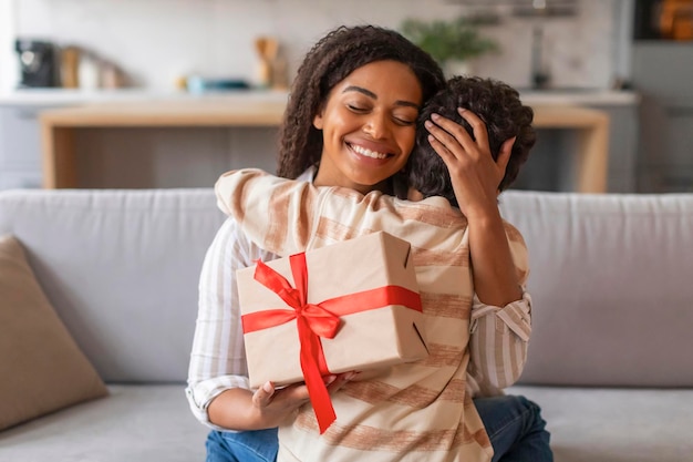 Радостная черная мать держит завернутую подарочную коробку и нежно обнимает сына