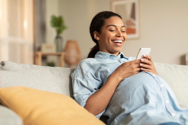 Joyful black pregnant woman using smartphone while relaxing on couch watching funny videos and laughing copy space