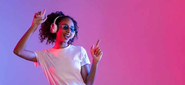 Joyful black woman with sunglasses and headphones dancing on neon background free space