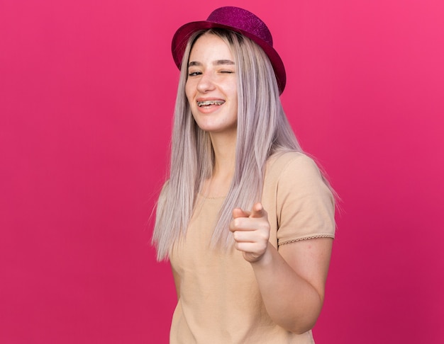 Joyful blinked young beautiful girl wearing party hat with dental braces showing you gesture 