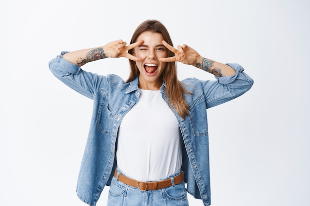 Gioiosa modella bionda dall'aspetto gioioso con segni a v sugli occhi, divertirsi e godersi la vita, in piedi positivo contro il muro bianco in camicia di jeans casual e t-shirt