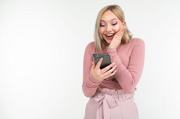 Joyful blond girl in pink stylish clothes in surprise rejoices at winning on the Internet by glancing into a smartphone on a white background with copy space