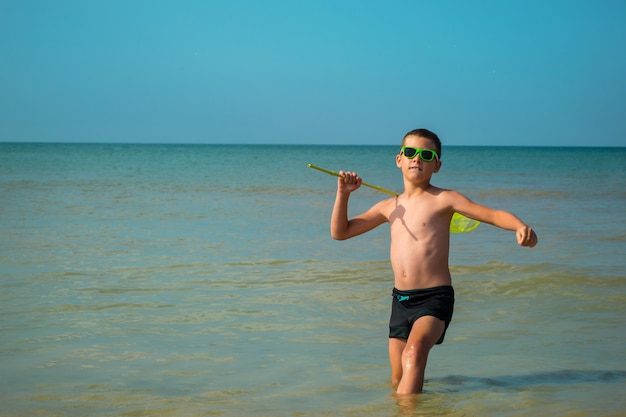 Joyful boy in sunglasses runs into the sea with a butterfly net.