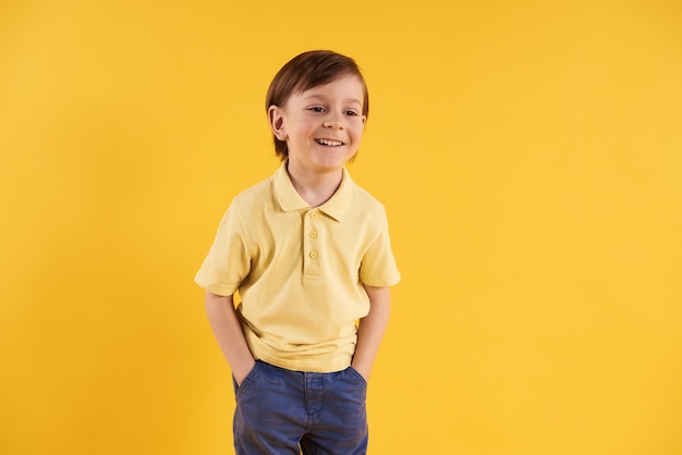 Joyful boy with hands in pockets on yellow background.