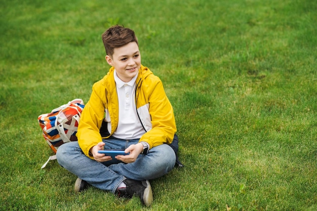Joyful boy in a yellow jacket sits on a green lawn with a smartphone in his hands