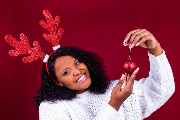 Joyful brazil charming woman in toy deer horns holding red ball gift box while at cozy room