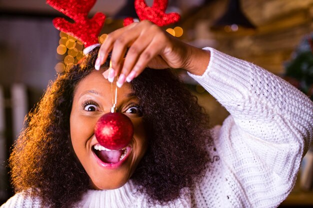 Joyful brazil charming woman in toy deer horns holding red ball gift box while at cozy room