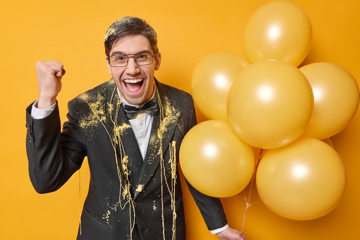 Joyful brunet man clenches fist exclaims from joy dressed formally smeared with serpantine spray holds bunch of inflated balloons isolated over yellow background People and celebration concept