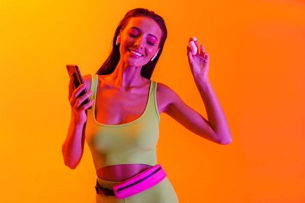  joyful brunette woman wearing formfitting sportswear dancing while using smartphone and earpods isolated over orange neon wall