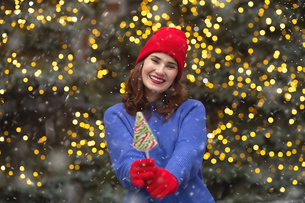 Joyful brunette woman with candy at the christmas tree during the snowfall