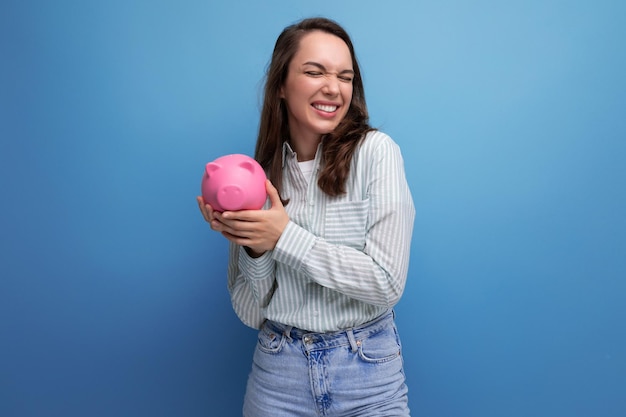 Joyful brunette young business woman in a shirt with a piggy bank on the background with copy pace