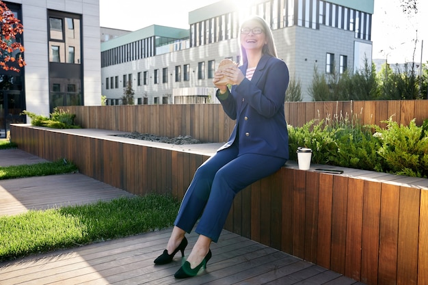 Joyful business-lady laughing and holding delicious burger while sitting on terrace and having her lucnh break