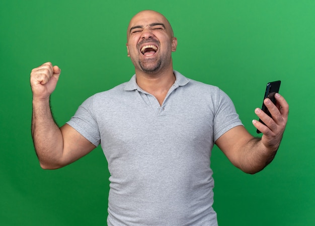 Joyful casual middle-aged man holding mobile phone doing yes gesture with closed eyes isolated on green wall