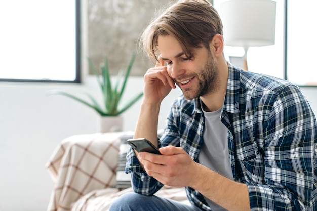 Joyful caucasian attractive guy using his smartphone while sitting on sofa at cozy home browsing internet and social networks texting with friends or family found out good news smiles happily