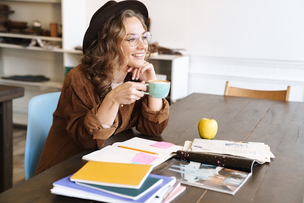 joyful charming woman in eyeglasses drinking coffee while studying with exercise books at home