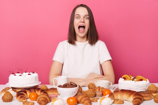 Joyful cheerful brown haired woman in white t shirt sitting at table i with pastry and different sweets yelling with joy ending diet isolated over pink background