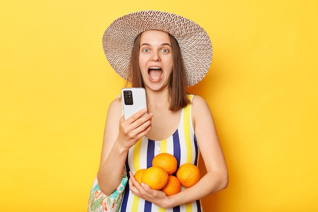 Joyful cheerful unhappy woman wearing striped swimming suit and hat isolated on yellow background using mobile phone reading news rejoicing expressing happiness