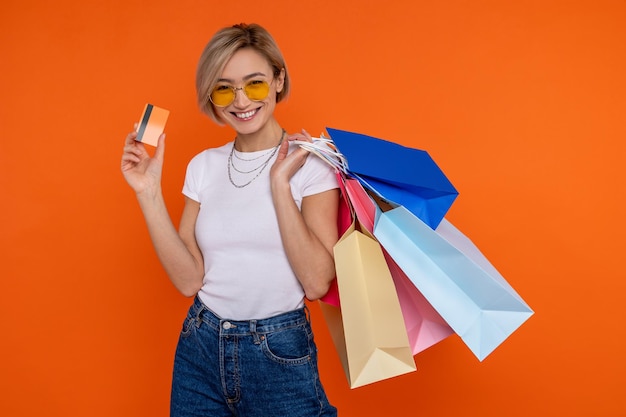 Photo joyful cheerful woman in white t shirt and jeans showing credit card and her purchases isolated over orange background