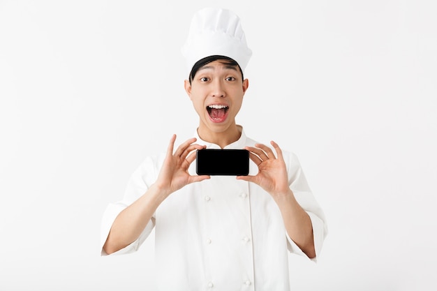 Gioioso capo uomo cinese in uniforme bianca da cuoco e cappello da cuoco che tiene il telefono cellulare isolato sopra il muro bianco