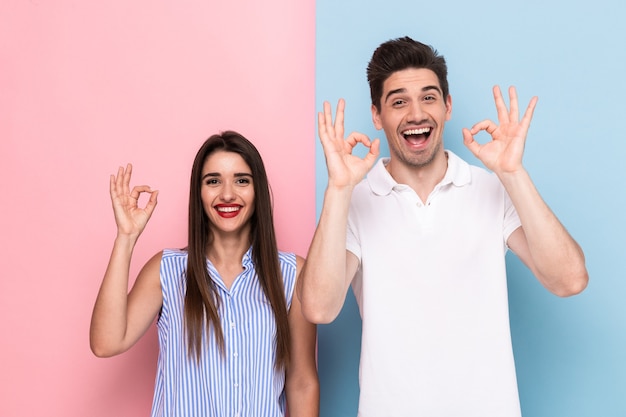 joyful couple in casual wear smiling and gesturing ok sign, isolated over colorful wall